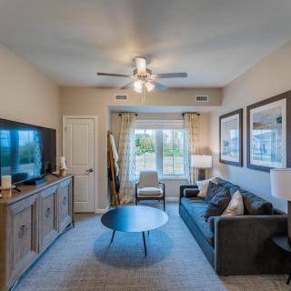 A cozy living room with a sofa, coffee table, and television, featuring a large window with curtains and a ceiling fan.
