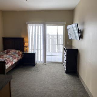 A cozy bedroom with a single bed, wall-mounted television, dresser, and sliding glass door allowing natural light in.