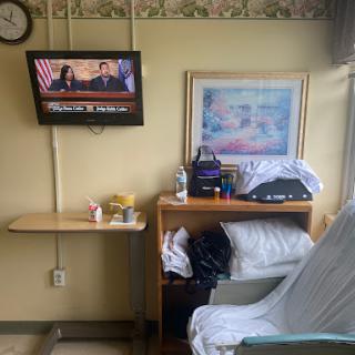 A view of a hospital patient room featuring a television, a bed, and some personal items.