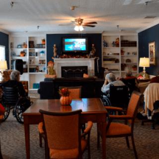 A cozy living room with seniors seated in wheelchairs watching television near a fireplace.