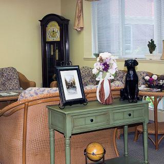 A cozy living room featuring a grandfather clock, wicker sofa, and a table with decorative items like a vase of flowers and a photo frame.