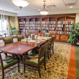 A cozy library room with a large wooden table surrounded by chairs, bookshelves filled with books in the background, and plants for decoration.