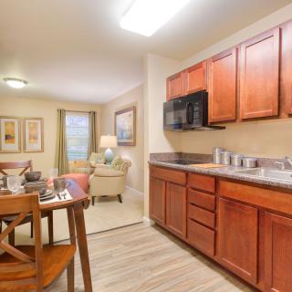 A cozy apartment interior featuring a small kitchen with wooden cabinets, a dining table, and a living room with warm lighting.