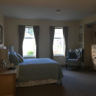 A cozy bedroom featuring a double bed, two windows with curtains, a comfortable chair, and a dresser.