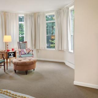 A cozy living room with an armchair, ottoman, and a lamp by large windows with curtains.