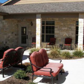 Outdoor patio area with cushioned chairs and landscaping in front of a brick building.