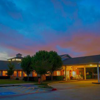 A warmly lit building with a parking area in the foreground, set against a colorful, cloudy evening sky.