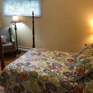 A cozy bedroom with a colorful bedspread, featuring a lamp and a comfortable chair by the window.