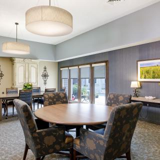 A well-lit dining room with a round table and upholstered chairs, featuring large windows and elegant lighting.