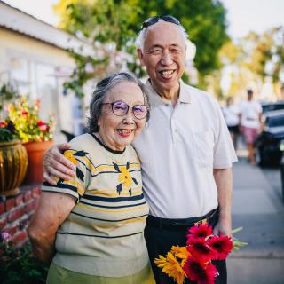 An elderly couple smiling and holding flowers while standing outdoors.