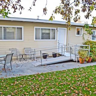 A house with a ramp leading to the entrance, surrounded by a patio with chairs and autumn leaves on the grass.