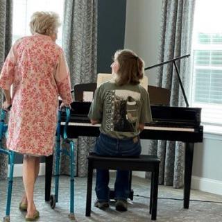 A child playing the piano while an elderly person stands nearby with a walker.