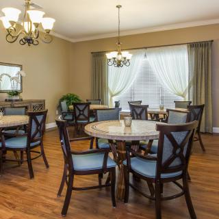A well-furnished dining room featuring round tables, cushioned chairs, and elegant chandeliers with ample natural lighting.