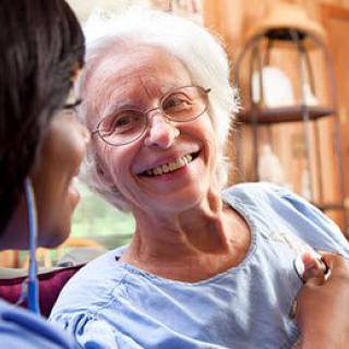 An elderly woman smiling while engaging in a conversation with her caregiver indoors.