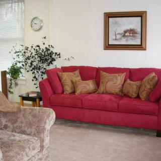 A cozy living room featuring a red sofa with decorative cushions, an armchair, and a potted plant. A clock and framed artwork adorn the walls.