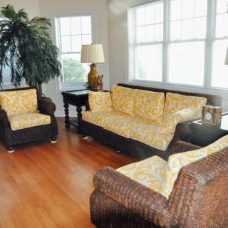 A cozy living room featuring wicker furniture, a large indoor plant, and abundant natural light from tall windows.