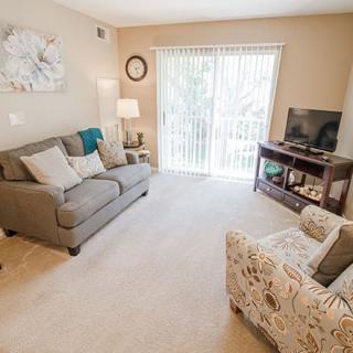 A cozy living room with a grey sofa, patterned armchair, TV on a stand, and soft lighting.