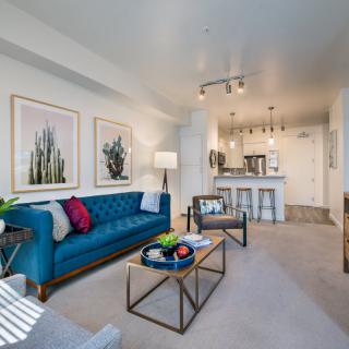 A modern living room with a teal sofa and contemporary decor, featuring a coffee table and artwork, leading into an open kitchen.