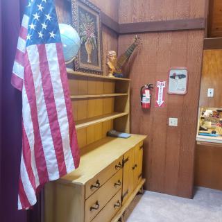 A room with an American flag, a dresser and shelves, featuring wood paneling and a fire extinguisher.