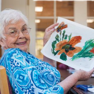 An elderly woman proudly displays her floral painting indoors.