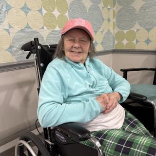 A cheerful senior woman in a wheelchair, wearing a pink hat and light blue top, smiles warmly.