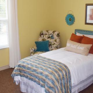 A cozy bedroom with a single bed, decorative pillows, and an armchair beside a window.