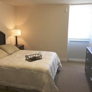 A cozy and simple bedroom featuring a bed, lamp, and dresser with natural light coming through the window.