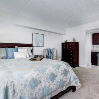 A cozy bedroom with elegant bedding and furniture, featuring a bed, dresser, and a small kitchenette area.