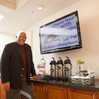 A man standing by a coffee station with a television displaying event details.