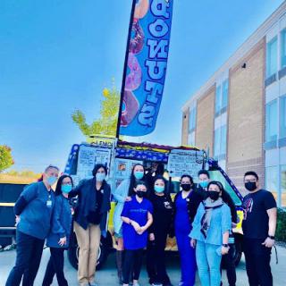 A lively group gathers in front of a colorful donut food truck at a community event.