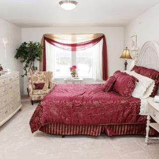 A cozy bedroom with red and white decor, featuring a comfortable bed, armchair, and a window with drapes.