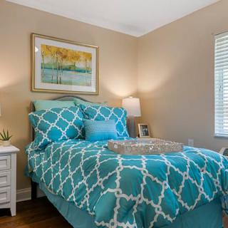 A cozy bedroom with teal patterned bedding, illuminated by warm lamp light, and featuring decorative artwork above the bed.