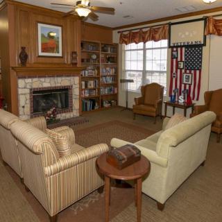 A cozy living room with a fireplace, couches, and armchairs. A bookshelf is in the corner, and a flag is displayed on the wall.