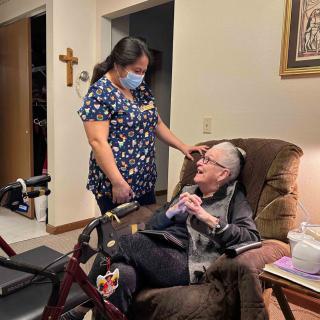 A caregiver wearing a mask interacts with an elderly person seated in a chair.