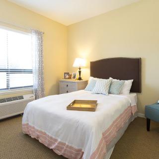 A cozy bedroom with a neatly made bed, a bedside lamp, and a window allowing natural light.
