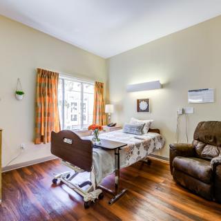 A well-lit hospital room with a patient bed and a recliner chair, featuring wooden floors and a large window with curtains.