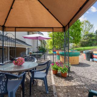 A cozy outdoor patio area with a gazebo, table, and chairs, surrounded by greenery and potted plants on a sunny day.