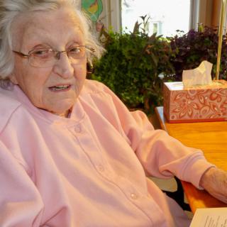 An elderly woman wearing glasses and a pink sweater is sitting at a table indoors.