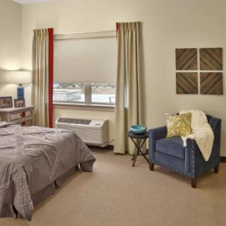 A cozy bedroom featuring a bed, armchair, and window with curtains.