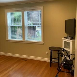A small room with wooden flooring, featuring a window with blinds, a television mounted on the wall, a stool, and a walker in the corner.