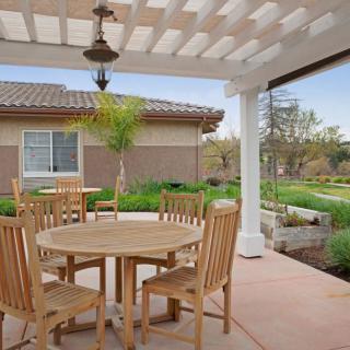A cozy patio area with a wooden table and chairs under a pergola, surrounded by lush garden landscaping.