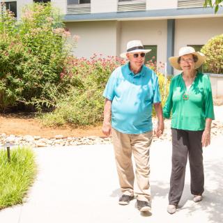 An elderly couple walks together in a sunny garden, both wearing hats.