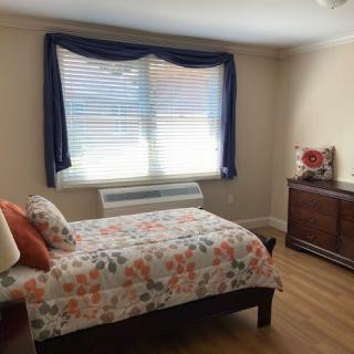 A cozy bedroom with a single bed and floral bedding, featuring a large window with curtains and a wooden dresser.