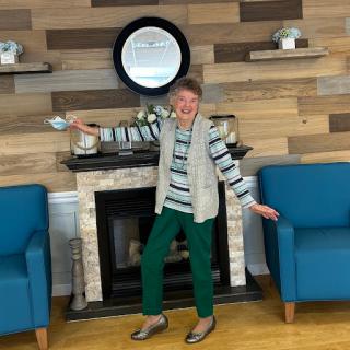 An elderly woman joyfully poses in a cozy living space with stylish blue chairs and a decorative fireplace.