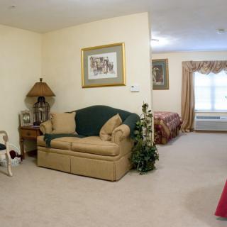 A cozy living room with a sofa and a lamp, leading into a bedroom with natural light.