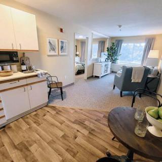 A cozy apartment interior featuring a kitchenette, living area, and large windows letting in natural light.
