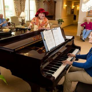 An elderly man plays the piano for an audience in a cozy living room.