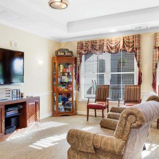 A cozy living room with a television, bookshelf, sofa, and chairs near a large window with curtains.