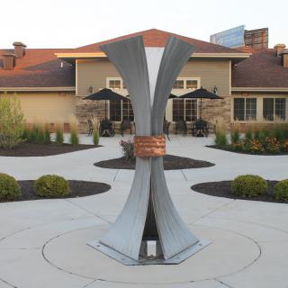 A modern metal sculpture stands in an outdoor courtyard, accentuated by surrounding landscaping and a building in the background.