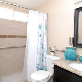 A modern bathroom featuring a shower with a floral curtain, toilet, countertop with sink, and mirror. Decor includes turquoise accents.